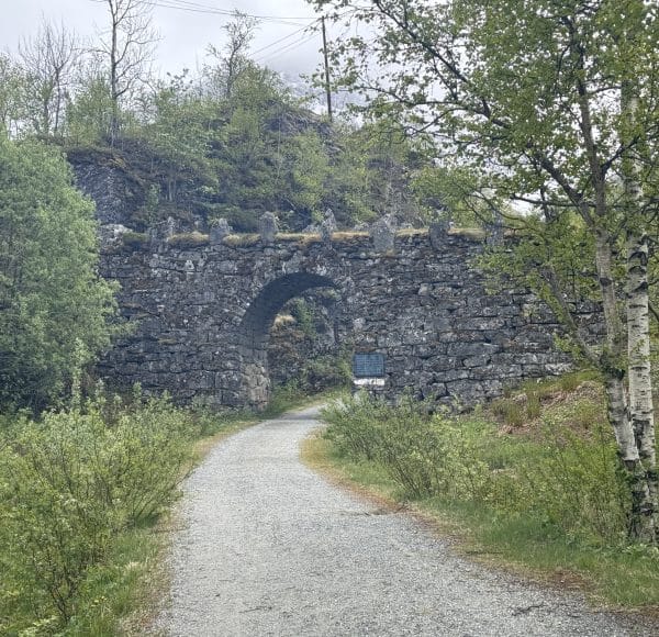 Steinerner Torbogen auf einem bewaldeten Wanderweg, umgeben von grüner Vegetation und Bäumen an einem bewölkten Tag.