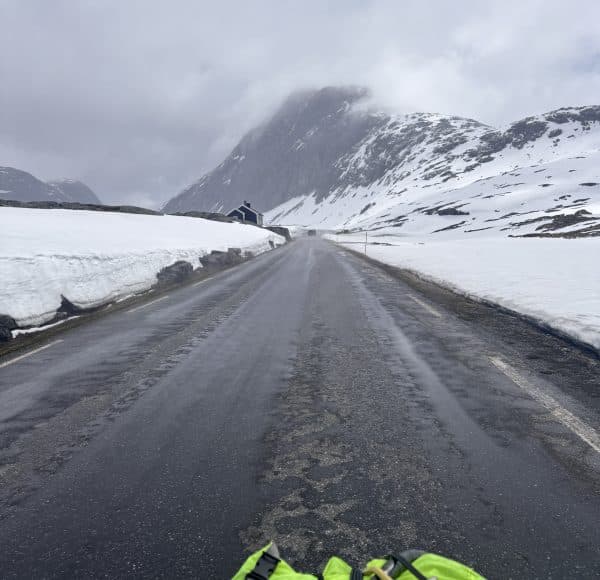 Nasse Straße durch schneebedeckte Berge, bewölkter Himmel, Haus am Straßenrand, vorne Teile einer grünen Tasche sichtbar.