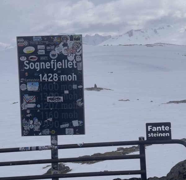 Verschneite Berglandschaft mit einem Schild „Sognefjellet 1428 moh“, beklebt mit Aufklebern, und einem kleineren Schild „Fantensteinen“, umgeben von Schneeflächen und Bergen im Hintergrund.