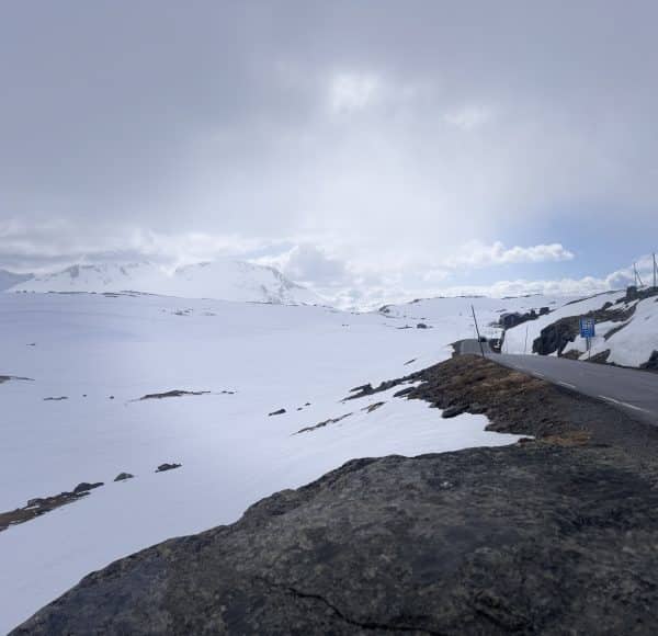Verschneite Berglandschaft mit vereinzelt sichtbaren Felsen am Straßenrand und teils bewölktem Himmel, Abschnitt des teilweise kurvigen Sognefjellsveien, höchster Pass Nordeuropas.