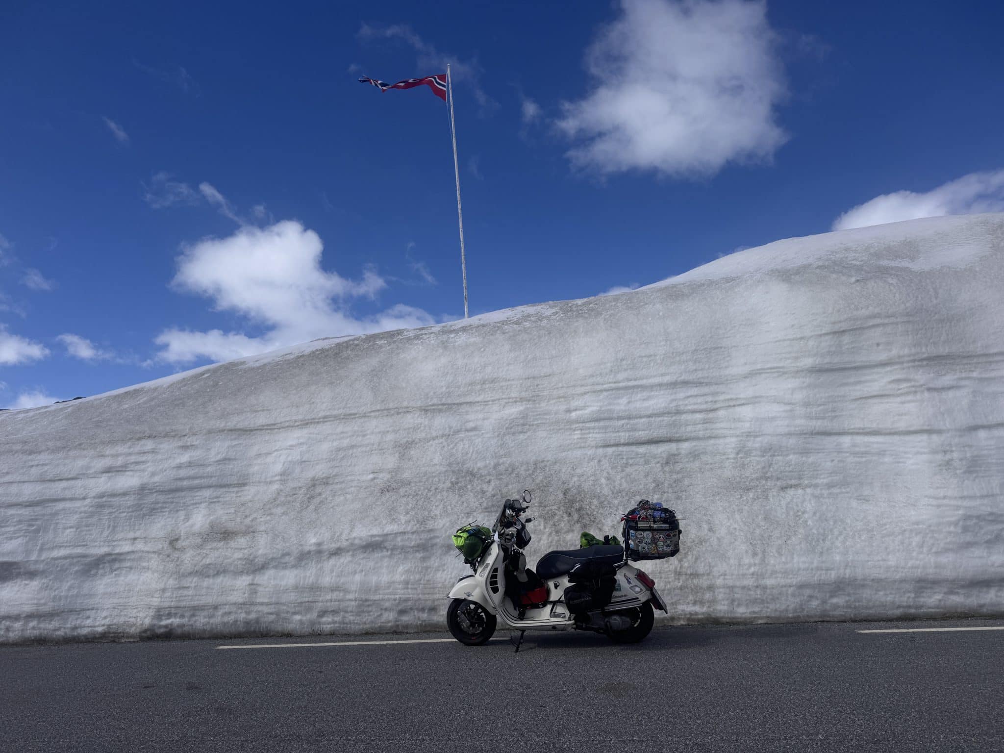Motorrad steht auf Straße vor hoher Schneewand, darüber weht eine norwegische Flagge, blauer Himmel mit wenigen Wolken im Hintergrund.