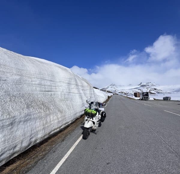 Motorrad steht auf einer Straße neben einer hohen Schneewand, im Hintergrund Berge und blauer Himmel mit wenigen Wolken.