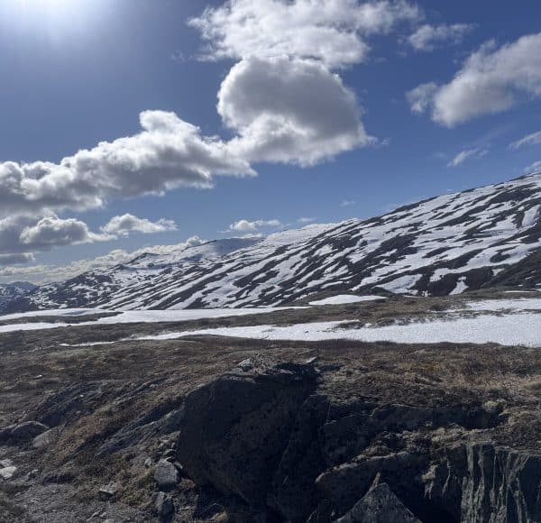 Sonniger Tag in den Bergen, schneebedeckte Hänge, teilweise freiliegende Felsen und Wiesen, dramatische Wolken am blauen Himmel.