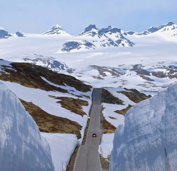 Sognefjellsveien (Fv 55) mit hohen Schneewänden führt durch verschneite norwegische Berge unter blauem Himmel; ein Auto fährt die kurvige Strecke entlang. © JustKommunikation GmbH | JustRide it