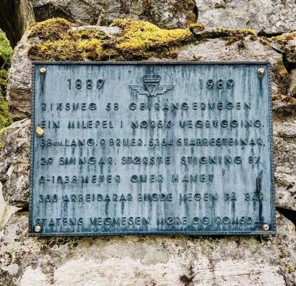 Metalltafel mit norwegischem Text über den Geirangerweg auf einem Felsen, umgeben von Moos und Felsenlandschaft.