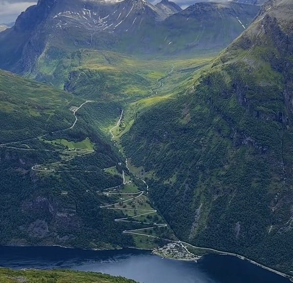 Verschlungene Serpentinenstraße führt durch grünes Tal, umgeben von hohen Bergen mit Restschnee, im Vordergrund ein ruhiger See und kleines Dorf am Ufer.