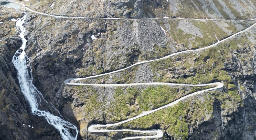 Atemberaubende Serpentinen mit einem Wasserfall, der steile Felsen hinabstürzt - Trollstigen aus der Vogelperspektive fotografiert.